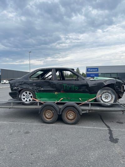 A black sedan, heavily modified with a roll cage and significant body damage, sits on a trailer. Green paint is applied in broad strokes along the lower body of the car, and the word "Tiril" is written in green with a heart symbol on the driver's side door. The car is parked on asphalt, with a warehouse-like building in the background bearing the "thansen" logo. The sky is overcast. The scene is located in Slitu, Norway.