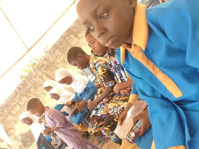 The image depicts a group of approximately six to seven individuals, primarily children and young adults, engaged in a focused activity, likely a lesson or religious instruction, in Ijoko, Nigeria. They are arranged in a diagonal line from the foreground to the background, generally looking downwards.
In the immediate foreground, a dark-skinned male youth, with a bald head, is visible from the chest up, wearing a bright blue long-sleeved garment with a yellow spread collar and a yellow fabric strip running down the front. He holds a rectangular book with a light-colored, possibly metallic or reflective, cover in his hands, which are positioned in his lap. His gaze is directed downwards and slightly to his right.
Behind him, another male youth wears a light purple long-sleeved tunic and also appears to be focused downwards. Following him are several females, most wearing white hijabs that cover their heads and necks, with varying dresses underneath. One wears a blue dress, another a multi-colored patterned dress with floral or abstract designs. They too are looking downwards, and at least one is seen holding a dark-colored book or object.
The background features a light-colored concrete block wall, partially obscured by bright sunlight from above, suggesting an outdoor or semi-outdoor setting under a canopy or overhang, indicated by white structures in the upper left. Green foliage is faintly visible behind the wall. The overall impression is one of students or participants in a structured learning environment.