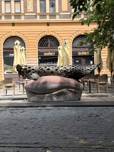 The image depicts an outdoor scene in Debrecen, Hungary. In the foreground, a sculpture made of dark metal and reddish-brown material is prominently displayed on a cobblestone street. The sculpture is abstract, featuring a textured, rounded base topped with a lattice-like structure. Behind the sculpture, a building with an orange facade features arched windows with ornate grilles. Two of these windows house businesses with signs that read "HONEY BAR & CLUB". Yellow umbrellas are set up in front of these establishments, and stacks of empty chairs are visible on the left side. The upper portion of the building shows more arched windows and decorative architectural elements. Green foliage from a tree hangs into the top right corner of the frame, suggesting an urban setting with some greenery. The overall scene appears to be a quiet daytime view of a street with an outdoor seating area for a bar or club.