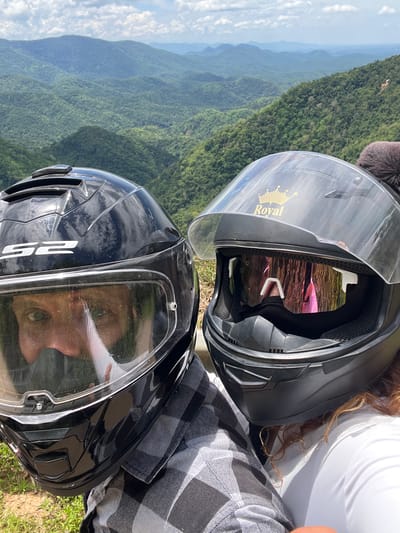 A selfie taken by two individuals wearing motorcycle helmets is captured against a backdrop of lush, verdant mountains under a cloudy sky. The person on the left, wearing a black helmet with "LS2" branding, has their eyes visible through the visor. The person on the right wears a black helmet with a gold "Royal" crown decal on the top and mirrored goggles. The helmets obscure the faces of the individuals, with only the eyes and parts of their faces visible. The background shows a panoramic view of rolling green hills and mountains, characteristic of a mountainous region like Son Dien, Vietnam. The lighting suggests it is daytime.