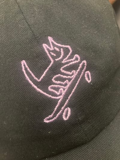 The image shows a close-up, angled view of a black baseball cap with a purple embroidered design. The design appears to be a stylized animal or character. The hat is made of a twill fabric with visible texture and stitching. There are small white specks, possibly lint or dust, scattered across the surface of the hat. The lighting is somewhat uneven, creating subtle shadows. The specific location in Cerdanyola del Vallès, Spain, is not discernible from the image itself.