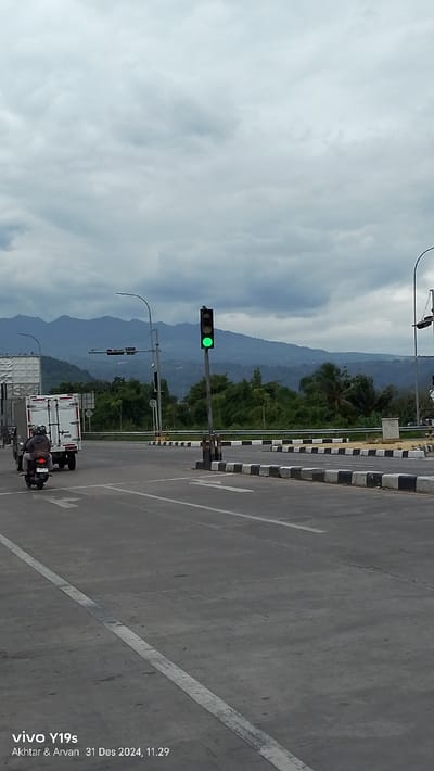 This vertical shot depicts a road scene in Sumedang, Indonesia, under a heavily overcast sky. The time of day is late morning, specifically 11:29 AM on December 31, 2024, as indicated by the timestamp.
In the background, a range of large, dark mountains covered in dense green foliage stretches across the horizon, appearing hazy under the cloudy conditions. The sky above is filled with thick, grey clouds, suggesting a muted light or potentially impending rain.
The foreground and midground are dominated by a paved road, likely an intersection or a turning point. A tall traffic light pole stands prominently on the right side of the road, displaying a bright green signal, indicating that vehicles can proceed. White road markings, including straight and curved directional arrows, are painted on the grey asphalt surface. A median strip, clearly demarcated with alternating black and white stripes, separates different sections of the road.
On the left side of the frame, a white box truck is partially visible, moving forward. Immediately behind it, a person wearing a dark jacket and a helmet is riding a motorcycle, also heading in the same direction. Other street furniture, such as light poles and a small electrical box, are visible along the median and roadside. The overall scene captures a moment of typical traffic flow within a landscape that blends urban infrastructure with natural mountainous beauty.
A digital watermark in the bottom-left corner reads "vivo Y19s Akhtar & Arvan 31 Des 2024, 11.29".