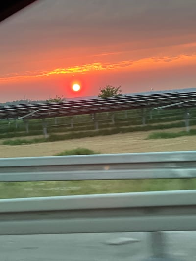 The image shows a vivid sunset with orange and red hues, as viewed through a window. The window frame is prominent in the foreground with multiple vertical bars. Outside the window, a rural or agricultural landscape is visible, with fencing and vegetation. A small tree or bush is present in the mid-ground. The overall scene captures a natural setting at sunset. Location information, including the city of Bedburg, Germany, cannot be confirmed from the image.