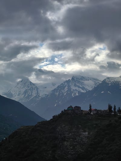 This is a photograph of a village nestled on a hillside with a backdrop of snow-capped mountains. The sky is heavily clouded, with patches of blue sky visible through breaks in the clouds. In the foreground, on the crest of a dark, tree-covered hill, a cluster of buildings forms the village. The structures appear to be of various sizes and architectural styles, with some featuring multi-story designs and pitched roofs. Several tall, slender trees are interspersed among the buildings. The midground is dominated by the imposing mountains, their peaks dusted with snow and their slopes a mix of rock and sparse vegetation. The overall impression is one of a remote and rugged landscape, likely in a mountainous region such as the Himalayas. The village is identified as Kardang, India.