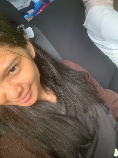 A young woman with dark hair is reclining. She has light brown skin and dark eyes. She is wearing a dark-colored top over a brown sweater or jacket. Some of her hair is spread out around her face. In the background, there is a light-colored garment, a dark grey surface, and a hint of blue and red cloth or materials. The location within the city of Samarinda, Indonesia, cannot be determined from the image.