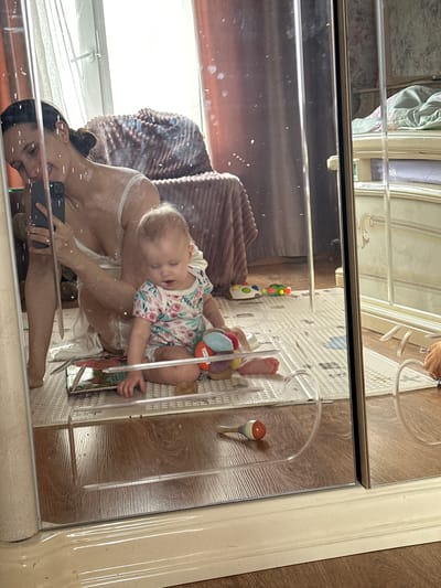 A person holds a baby in front of a mirror, taking a photo with a cell phone. The baby is wearing a floral shirt and is asleep or drowsy. A colorful toy is near the baby. The mirror reflects a bed and window. A play mat and a fishing bobber are visible near the baby. The location is likely inside a home. There is no indication that the location is in the city of Pokrov, Russia.