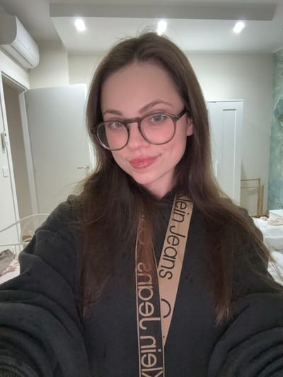 The image presents a headshot selfie of a young adult female indoors. She possesses fair skin, light-colored eyes, and long, straight dark brown hair, parted slightly off-center. She wears dark-rimmed, round spectacles and a dark-colored hooded sweatshirt. A beige strap with the text "Calvin Klein Jeans" printed repeatedly in black lettering is visible diagonally across her chest. She is smiling faintly with closed lips, directly engaging with the camera.

The background depicts a light-colored room. The ceiling is white and features two recessed, circular light fixtures. On the upper left, a white wall-mounted air conditioning unit is visible. Below it, a white door frame leads to a closed white door with a silver handle. A light switch or control panel is mounted on the wall to the left of this door. On the right side of the image, another closed white door is visible. Behind the subject's left side, a section of wall exhibits a light blue/green and white patterned or textured finish, possibly a mural or wallpaper. Below this, a light-colored textile, suggestive of bedding, is present. A light-colored wooden frame structure is partially visible near the floor on the right. There are no visual indicators within the image to confirm the location as Scafati, Italy.