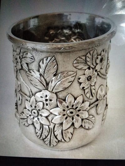 A silver cup with an ornate floral design sits on a flat surface. The cup's exterior is adorned with detailed embossed flowers and leaves, giving it a textured appearance. The interior of the cup is dark and appears to contain some reflective material. The background is light and slightly out of focus. There is no indication of any specific location in the city of Kualaraja, Indonesia, within the image itself.