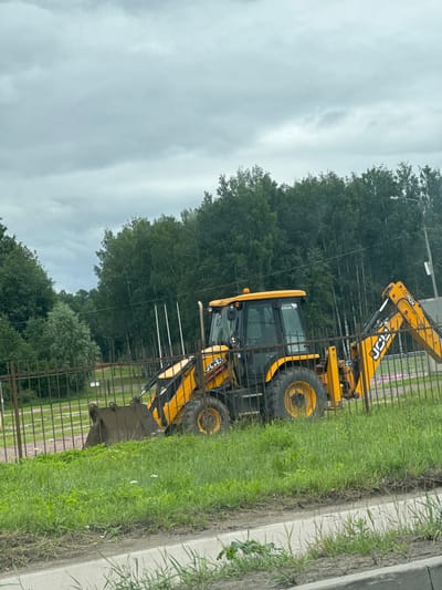An orange and yellow JCB backhoe loader is parked on a grassy area behind a metal fence. The backhoe is positioned with its front loader bucket lowered to the ground and its rear digging arm extended slightly. The machine has large black tires and an orange flashing light on its roof. The cab of the backhoe has tinted windows. A sticker on the side of the cab reads "СДЕЛАНО В РОССИИ" (Made in Russia). The scene is framed by a chain-link fence, with tall green trees forming a dense backdrop beyond the fence. The sky is overcast with gray clouds. The ground in front of the fence is covered in grass with scattered white wildflowers. A paved surface is visible in the foreground. The overall environment suggests an outdoor location, possibly a construction site or a vacant lot within the city of Pargolovo, Russia.