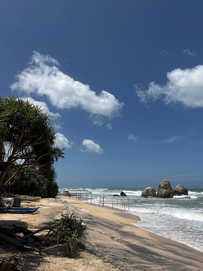 The image depicts a serene beach scene in Habaraduwa, Sri Lanka, under a clear blue sky with scattered white clouds. The foreground features a sandy beach with some scattered debris from trees and a few low-lying green plants. Two small fishing boats are pulled up onto the sand, one predominantly blue and red, and another lighter colored one behind it.
The midground shows the transition to the ocean with gentle waves lapping at the shore. A series of thin, dark poles are partially submerged in the water, extending from the beach into the sea, likely part of a fishing setup. Further out, several large, grey rocks are situated in the water, with waves crashing against them.
The background reveals the vast ocean and the horizon where the sea meets the sky. On the left side of the image, lush green vegetation, including trees with broad leaves and branches, grows along the edge of the beach, providing shade. The overall impression is one of a peaceful, natural coastal environment.