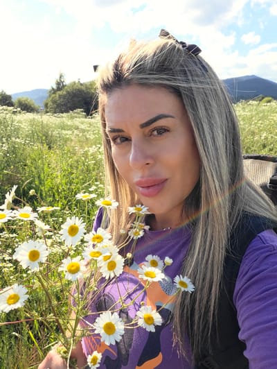 A woman with blonde hair styled in loose waves is taking a selfie outdoors in a field of daisies. She is wearing a purple t-shirt with an orange and black graphic design. She is holding a bouquet of daisies in her left hand. The background shows a meadow filled with white daisies and green grass, with trees and mountains in the distance. The sky is blue with some white clouds. The location is the village of Ravnogor, Bulgaria.