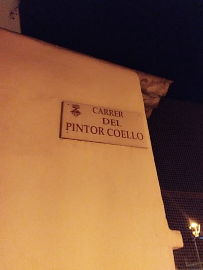 A street sign on a corner building at night reads "Carrer del Pintor Coello" in Rubí, Spain. The building is light yellow or tan in color. A streetlight is visible behind a wire fence.