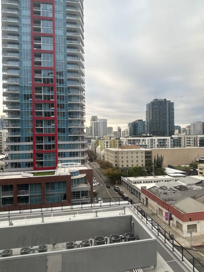 An urban landscape features a tall building with red and blue windows dominating the left side of the frame. Further in the distance are other buildings and skyscrapers, with a cloudy sky above. To the right is a section of a building with visible fans and rooftops of nearby structures. A street with parked cars is visible between buildings. The scene is oriented vertically.