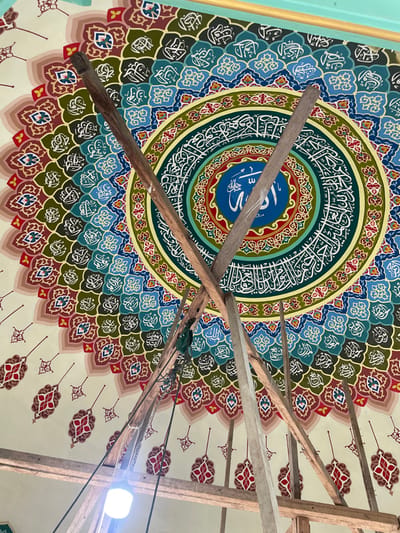 The image depicts the interior of a building, presumably a mosque, located in Kumbang, Indonesia. The central subject is a circular, richly decorated dome ceiling painted with Islamic calligraphy and intricate geometric and floral patterns.

The dome's center features a bright blue medallion with white Arabic script, presumably "Allah," surrounded by concentric rings. These rings display further white Arabic calligraphy against various colored backgrounds (teal, dark green) and repeating patterns of stylized motifs in shades of red, blue, green, and brown. The outermost visible sections of the dome display individual, elongated, red and cream colored motifs against a light cream background, extending towards a light teal border at the top edge of the frame.

In the foreground, a structure of rough-hewn wooden poles forms scaffolding, partially obscuring the dome's painted surface. Several poles are crisscrossed, and horizontal beams are visible below. A single, unshaded, bright white LED light bulb hangs from dark wires in the lower-left portion of the image, attached to the scaffolding structure. Green strapping or rope is also visible near the scaffolding. No people are present in the scene. The overall environment suggests ongoing construction, renovation, or maintenance within the building.