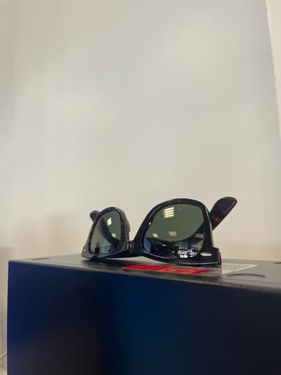 A pair of tortoiseshell-patterned sunglasses with dark green lenses rests on a dark blue rectangular box in the city of Gandra, Portugal. The sunglasses are positioned horizontally, slightly angled with one temple arm partially extended upwards and backward to the right. Reflections of square grid overhead fluorescent lights are prominent on both lenses. A partial red label or sticker is visible on the box surface beneath the sunglasses. The background is a plain, uniformly lit white wall. The scene is static and devoid of human presence or overt action.