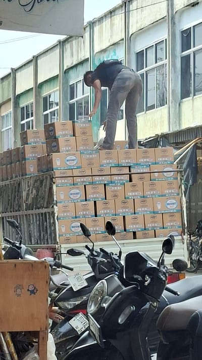 A man in a black t-shirt and grey pants is standing on top of a truck filled with stacked boxes, leaning over to reach something. The boxes are labeled with a blue logo featuring a cup. The truck is parked in front of a building with a pale green facade and numerous windows. In the foreground, several motorcycles are parked, with one prominent wooden crate. The scene appears to be outdoors, possibly in a market or commercial area, given the presence of the truck and motorcycles. The image is taken from a slightly elevated angle, looking up towards the man on the truck.