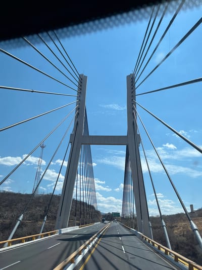 The image shows the Mezcala Bridge, a cable-stayed bridge located in Tula del Río, Mexico. The view is taken from a vehicle on the bridge deck looking upwards at the towers and supporting cables. The bridge structure is characterized by its two central towers connected by a horizontal beam at the top. The cables radiate outward from the towers to support the bridge deck. The sky is blue with scattered clouds. Adjacent to the bridge is a steep, arid hillside. Part of a radio communication tower is visible behind one of the bridge towers. The bridge has an asphalt road surface with white and yellow lane markings. The perspective indicates the photograph was taken while in transit on the bridge.