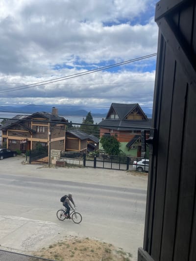 The image presents a daytime view looking down from a vantage point, likely a balcony or window, which frames the scene with dark wooden planks at the top and partially along the bottom right. The perspective looks out over a street and residential area with mountains in the background.

In the lower right quadrant of the image, a person is seen riding a bicycle on a light gray paved road. The cyclist is wearing a light-colored helmet, a dark long-sleeved top or jacket, and a dark backpack. They are facing away from the viewer, riding towards the left. The bicycle has a dark frame and wheels. To the immediate right of the road, there is a dry, grassy patch of land.

Across the street, in the midground, several houses with a distinct rustic architectural style are visible. These buildings feature a combination of dark wood siding and lighter-colored stone or brick accents, topped with steep, dark-pitched roofs. The house on the mid-left side of the frame has a sign near its entrance, though only part of the text is clearly legible, appearing to read "El [obscured word, possibly 'Puesto']". A dark sedan car is parked on the street in front of this particular house. Further back and slightly to the right, another large wooden house with a prominent A-frame or gabled roof stands. Various fences, green shrubs, and a few trees are scattered throughout the properties. Utility poles and dark power lines run horizontally across the middle of the frame, extending across the sky and in front of the distant houses.

In the far background, a range of large mountains rises, appearing somewhat muted or hazy in the distance. The sky above is partly cloudy, featuring patches of bright blue interspersed with white, fluffy cumulus clouds, suggesting a pleasant daytime setting. The scene evokes a tranquil, residential atmosphere within a mountainous or scenic natural environment.