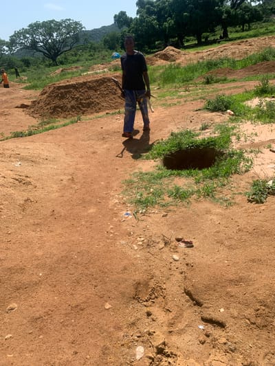 In Buga da Gindi, Nigeria, a young individual stands in a dusty, open area. They are wearing a dark t-shirt and blue pants with a white pattern. The individual is holding a shovel with a wooden handle and a white end. The environment is characterized by piles of brown dirt, sparse green grass, and a clear sky. In the background, there are large trees and hills. To the left of the individual, another person in an orange shirt is visible, also near a pile of dirt. A hole is present in the ground to the right of the standing individual. There is scattered debris on the ground, including a white plastic bottle and what appears to be a discarded shoe. The ground is predominantly reddish-brown earth.