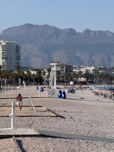 The image depicts a sunny day on a crowded beach in Pla Parcial Platja, Spain. The scene is dominated by a vast mountain range in the background, with a clear blue sky above. In the foreground, a person is engaged in some form of exercise on a beach apparatus. The beach itself is covered in pebbles, with numerous lounge chairs and umbrellas set up, indicating a popular tourist destination. Several buildings, likely hotels or apartments, line the edge of the beach, interspersed with palm trees. Blue bins are visible on the beach, and a life guard tower stands prominently. The overall atmosphere appears lively and recreational, suggesting a vacation or leisure activity. The time of day seems to be late morning or early afternoon, given the bright sunlight and shadows.