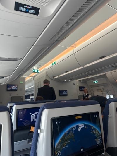 The image depicts the interior of an airplane cabin, viewed from the perspective of a passenger. In the foreground, several rows of airplane seats are visible, with their individual entertainment screens. The screen on the right displays a world map with various cities and locations highlighted. The screen on the left shows the Lufthansa logo and the text "28B". Above the seats, the overhead compartments and lighting fixtures are visible. In the middle ground, a flight attendant in uniform can be seen standing and talking to a passenger. Further back, another passenger in a dark suit is walking down the aisle. The lighting is a mix of overhead cabin lights and the warm glow of accent lighting along the ceiling. There are no discernible architectural elements suggesting a specific location within the city of Freising, Germany; the scene is entirely contained within the airplane.