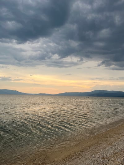 A person stands on a beach in Ofrynio, Greece, at what appears to be sunset. The sky is filled with dramatic, dark gray clouds, contrasting with the soft, golden light of the setting sun peeking through. The sea is calm, with gentle waves lapping at the shore. The beach itself is a mix of sand and pebbles. In the distance, a mountain range forms a silhouette against the horizon. The overall mood is one of tranquility and natural beauty.