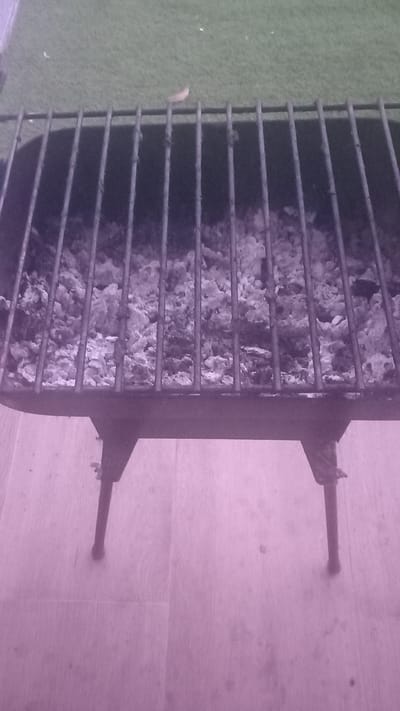 The image shows a close-up, rotated view of a barbecue grill. The grill is made of dark metal bars, and underneath the bars, there are remnants of ash and charcoal, indicating it has been used recently. The grill has two legs visible on the right side, propping it up. To the left of the grill, a patch of green, possibly artificial turf or a mat, is visible. The background consists of a light-colored wooden surface, likely a floor or tabletop, with faint grain patterns. The lighting in the image has a strong purple hue, suggesting artificial lighting or a color filter. No people are present in the image. The scene is likely outdoors or in a covered outdoor area given the presence of a grill and turf. There are no visible text elements in the image.