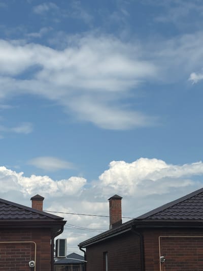 The image shows a view of the sky with clouds, above the rooftops of brick houses in the city of Krasnyi Krym, Russia. Two brick chimneys are visible extending from the dark brown tiled roofs. An outdoor air conditioning unit is mounted on the side of one of the brick houses. The sky is blue with scattered white cumulus clouds. The scene appears to be during daylight hours.