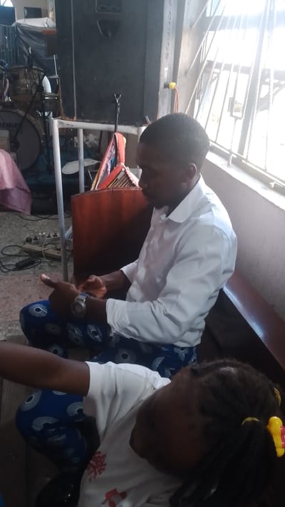 The image captures an indoor scene, likely within Warri, Nigeria, focusing on two individuals. In the foreground, a person is seen from the shoulders down, wearing a white t-shirt with a red graphic design and blue patterned trousers. Their hair is styled in braids and adorned with yellow and red hair accessories. To the left and slightly behind, an adult male is seated, wearing a white collared shirt and blue patterned trousers. He is looking down and appears to be interacting with his hands, one of which is adorned with a wristwatch. The background reveals a large black speaker and parts of a drum set, suggesting a music or performance-related setting. The overall environment appears to be dimly lit, with light filtering in from a window or opening on the right.