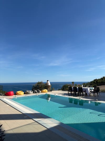 The image depicts an outdoor setting with a swimming pool and a view of the sea. A person, identified as a male, stands by a glass railing overlooking the water. In the foreground, a swimming pool with turquoise water is visible, with a tiled edge. Several beanbag chairs in red, yellow, and gray are placed near the pool. Further back, a dining table is set with a white tablecloth, chairs, and glassware. The environment suggests a residential or hospitality setting in Krimovice, Montenegro, characterized by its coastal landscape. The sky is clear blue with a few faint contrails.