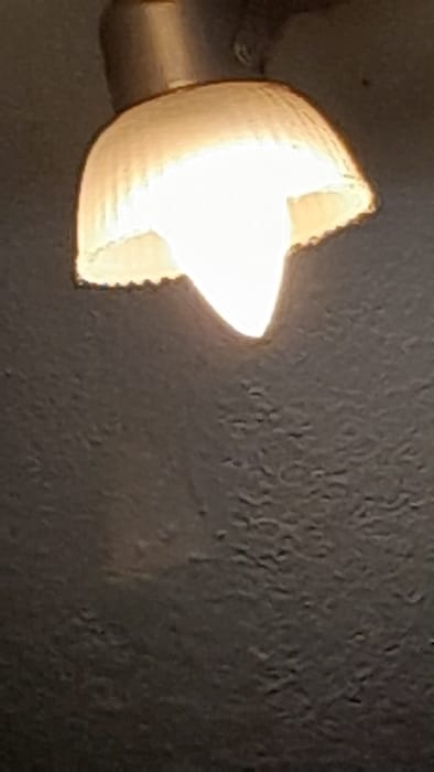 The image shows a close-up of a light fixture mounted to a wall. The light fixture is a white, off-white, or cream-colored dome with a broken, jagged edge. The light is on and the fixture is illuminating the surrounding area, which appears to be a wall or ceiling. The wall or ceiling is a dull, dark brown or gray color and appears to be textured. The location of the image is in the city of Merlo, Argentina, based on the provided coordinates. There is no indication of people or objects in the image besides the light fixture. The scene depicts a simple interior space, with no indication of activity, interaction, or specific time of day.