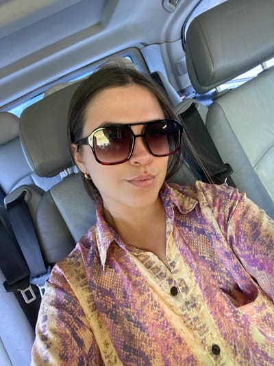 A woman with dark hair and sunglasses is taking a selfie in the back seat of a car. She is wearing a patterned shirt. The car interior appears to be gray leather. The image does not contain any information about the city of Leninkent, Russia.