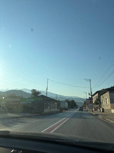 The image depicts a view from a vehicle driving down a street in Kresna, Bulgaria. The scene is bathed in the soft light of early morning or late afternoon. On the left side of the street, a building with a green sign that reads "Sava" is visible, with a wire mesh covering its storefront. Further down the street, a white truck is parked. On the right side of the street, a row of buildings lines the road. One building has a sign that says "SUPER SHOP." Several streetlights are present along the sidewalk. The road itself has a double white line with a red stripe in the middle, indicating it is a two-lane road. In the distance, rolling hills covered in greenery are visible against a clear blue sky. The overall impression is a quiet, rural or small-town street scene.