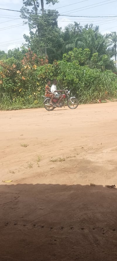 A medium shot captures two individuals on a red motorcycle traveling along a dirt road in Amauzu, Nigeria. The motorcycle rider, a male, is dressed in a white shirt and shorts. A female passenger is seated behind him, wearing a colorful dress. The road is flanked by dense green foliage and trees, typical of the region. Overhead, power lines stretch across the sky. The foreground is slightly obstructed by a shaded area, likely a building or structure from which the photograph was taken.