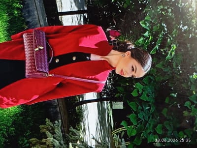 The image, taken in Griboyedov, Armenia, shows a woman wearing a red jacket and a black top. She has a burgundy clutch bag with gold hardware. The woman is positioned in an outdoor setting with lush green foliage surrounding her. The lighting suggests it is daytime. The timestamp "08.06.2026 23:35" is visible in the bottom right corner.