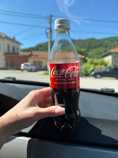 A partially full Coca-Cola bottle is held by a person's left hand, with their fingers wrapped around the lower portion of the bottle. The bottle has a red label with the Coca-Cola logo and text that reads "ORIGINAL TASTE FIFA WORLD CUP". The bottle is positioned in front of a car's dashboard, casting a shadow. The background, viewed through what appears to be a car window, shows a sunny outdoor scene with houses, trees, and a clear blue sky with some utility lines. The location is identified as Beli Osam, Bulgaria.