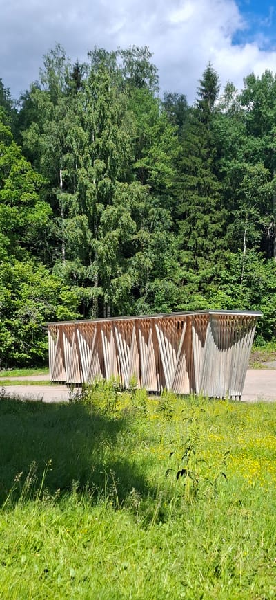 The image shows a modern wooden structure with a slatted facade, possibly a bus shelter or pavilion, situated in an outdoor setting. The structure is made of light-colored wood and has a sloped roof. It is positioned on a paved surface adjacent to a grassy area with wildflowers. Behind the structure, a dense forest of deciduous and coniferous trees forms the backdrop. The sky is partly cloudy with patches of blue. The scene is illuminated by natural daylight. The image does not contain any discernible people or specific city landmarks that would allow for precise location identification within Lahti, Finland, beyond it being an outdoor, natural environment.