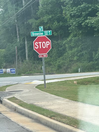The image shows a street scene in Mountain Park, Georgia, United States. A "Brownlee Road SW 5100" street sign and an "All Way" stop sign are prominently featured. Political campaign signs for "Matt & Venola Mason Judge" are visible alongside the road. The scene includes a sidewalk and greenery with trees in the background. The shot appears to be taken from inside a vehicle.