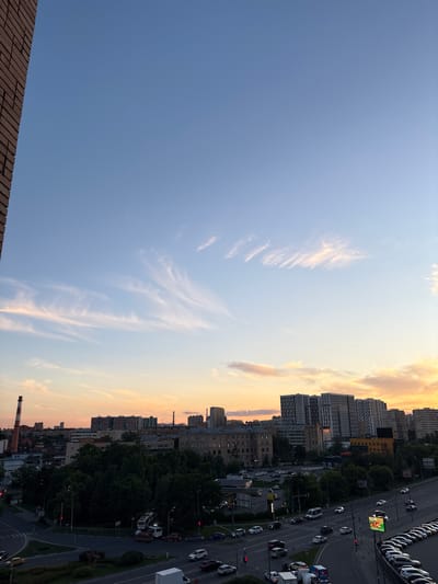 This is an aerial, high-angle shot of a city street at dusk. The sky is a mix of blue and orange, indicating sunset. There are several tall buildings and trees visible, with a busy street running through the middle of the frame. Cars and trucks are driving on the road, and there are a few pedestrians on the sidewalks. There is also a tall brick chimney in the foreground. The overall mood of the image is peaceful and serene, with a touch of urban energy.