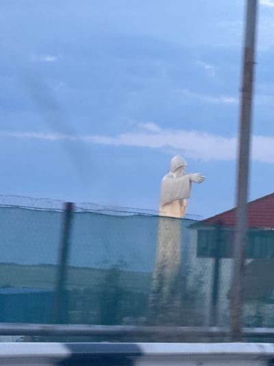 The image captures a statue of a robed figure with a hood, possibly depicting Jesus Christ, standing tall against a cloudy blue sky. The statue is positioned in the city of Yeghvard, Armenia, with its arms outstretched. In the foreground, a fence made of metal mesh is visible, partially obscuring the view. Behind the fence, a red-roofed building is partially visible. The overall scene suggests an outdoor environment, likely a public space or park, given the presence of the statue and the fence. The lighting indicates it might be late afternoon or early evening, with soft, diffused light.