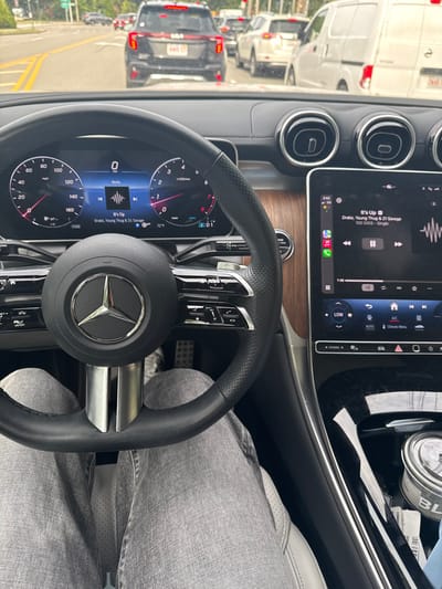 The image depicts a first-person view from the driver's seat of a Mercedes-Benz vehicle, showing the interior dashboard and a view through the windshield of a traffic scene in Newton, Massachusetts, United States.
**Interior Details:** The driver's legs, clad in grey pants, are visible below the black, multi-function leather steering wheel, which prominently features a chrome Mercedes-Benz logo. The digital instrument cluster displays "0 mph", indicating the vehicle is stationary, and shows "Media" playing "It's Up" by Drake, Young Thug & 21 Savage. The engine RPM gauge is at zero, and the ambient temperature is 72°F. To the right, a large vertical infotainment touchscreen reiterates the song title and artist, displaying a waveform visualization and "100 GIGS - Single," with the current time as 3:03. The screen also shows climate control functions including "LOW", "AUTO", "A/C", and "Climate Menu." The dashboard features a brown wood-grain trim element. The center console to the right includes a cupholder containing a metallic container and a blue object. Metallic pedals are visible on the floor.
**Exterior Details:** Through the windshield, a dense line of vehicles is observed in traffic on a paved road. Directly ahead is a black Kia Telluride SUV with a Massachusetts license plate reading "889 CXX." Further down the road, additional cars and a white cargo van are visible, all appearing to be stationary or moving slowly. The road features visible double yellow lines. Green foliage and trees line the roadside in the background, with a traffic light partially visible further in the distance.