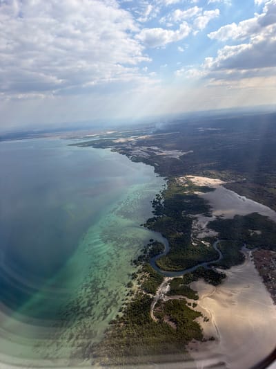 This is an aerial, landscape photograph taken from an airplane window. The image depicts a coastal scene, likely at low tide, showcasing a delta or estuary system with winding waterways and sandy banks juxtaposed against dense green vegetation. The water is a clear turquoise near the shore, becoming a deeper blue further out. Sunlight streams from the upper left, illuminating clouds and casting subtle shadows across the land and water. The scene is framed by a dark, curved edge of the airplane window. The time of day appears to be late morning or early afternoon, given the angle and intensity of the sunlight. The weather seems fair, with scattered white clouds in a bright blue sky. The location is identified as Metamelo, Mozambique, a region known for its coastal geography, which aligns with the visual evidence of river deltas and shallow, clear waters. There is no visible text in the image.