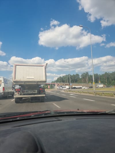 The image depicts a road scene in Sękocin Stary, Poland, under a bright, cloudy sky, suggesting it's daytime. The perspective is from inside a vehicle, looking out and slightly upwards, with a portion of the vehicle's interior visible on the right.

The main subjects are vehicles on a multi-lane road. In the foreground, a large white truck with a silver-colored trailer is prominent. The trailer has some writing on its side, but it's difficult to read clearly. The truck's license plate is visible and reads "WP 790AC". Behind this truck, the rear of a white van is visible, with partial text that appears to be "GS". Further down the road, other vehicles, including cars and trucks, are seen in traffic.

The setting includes a paved road with lane markings and a grassy verge. On the left side of the road, several tall streetlights are present, one of which is clearly visible in the foreground. Beyond the road, there is a dense line of trees, primarily pine trees, indicating a semi-rural or suburban area. Traffic signs are also visible along the roadside, including some yellow directional signs with black arrows.

The activity appears to be a traffic jam or slow-moving traffic on a highway or a major road. The bright sky and fluffy white clouds suggest fair weather. The overall mood is neutral, capturing a typical moment of travel.