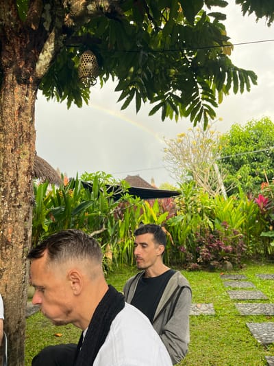 In Abiansemal, Indonesia, an outdoor scene unfolds under a cloudy sky with a visible rainbow. In the foreground, a man with a buzz cut and stubble is seen in profile, wearing a white shirt and a black scarf. His cheeks are puffed out, suggesting he might be eating. To his right, another man is seated, wearing a black t-shirt and a gray hooded jacket, looking to his left. A large tree trunk with rough bark stands to the left of the frame, its branches extending over the scene. Behind the men, a lush garden is filled with various green plants, some with red and yellow accents, and a pathway made of stepping stones. In the background, traditional Balinese structures with thatched roofs are visible amongst the foliage. A decorative woven light fixture hangs from a branch of the tree.