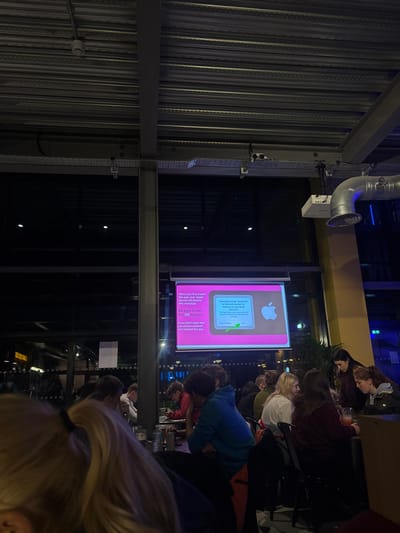 A low-angle indoor shot shows a group of people gathered near a display screen. The screen features an Apple logo and text about app installation and messages. The people are mostly facing the screen, some sitting at tables with drinks. The interior has exposed metal beams and ductwork, creating an industrial feel. The lighting is dim, with artificial light illuminating the screen and the surrounding area.
