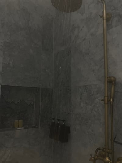 The image displays an interior view of a modern shower stall, characterized by a dimly lit environment. The walls are clad in large, rectangular tiles featuring a grey and white marble-like pattern with dark veining.

On the right side of the frame, a bronze-toned shower fixture system is visible. This system includes a prominent overhead rain showerhead, from which water is actively flowing downwards, creating multiple visible streams and splashes against the wet tile surface. Below the showerhead, a vertical pipe extends down to control valves and a wall-mounted holder for a bronze-toned handheld shower sprayer connected by a flexible hose.

On the left side of the shower stall, a recessed rectangular niche is cut into the tiled wall. Within this niche, two small, light-yellow colored objects, possibly soap bars or small bottles, are arranged on a white tray or base alongside a thin white object. Adjacent to the niche, on the wall facing the viewer, three dark-colored, uniform pump dispensers are mounted vertically.

No individuals, animals, or specific identifying marks for a precise geographical location are present within the image. The specific location within Santa Ana Portales, Mexico, cannot be determined from the visual information provided.