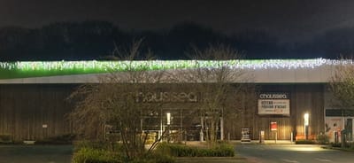 A Chausséa store at night, adorned with white and green Christmas lights along the roof. A large sign on the store displays "CHAUSSEA" and "DÉFEND LE POUVOIR D'ACHAT." The building has a wooden facade. Bare trees are in front, partially obscuring the entrance. The parking area is visible with a crosswalk in the foreground. Overall, the image has a dark, nighttime ambiance.