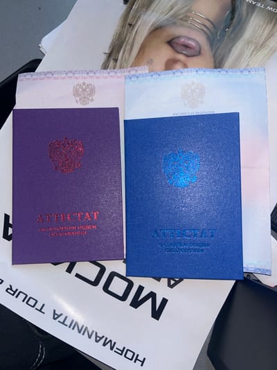 In Lobnya, Russia, a photograph captures a close-up view of two Russian "Attestat" documents, one purple and one blue, lying on a white paper with black text. The purple document, on the left, features a red embossed emblem of the Russian double-headed eagle and the embossed text "ATTESTAT" with smaller text below. The blue document, on the right, has a blue embossed double-headed eagle and embossed text "ATTESTAT" with smaller text beneath it. Both documents appear to be official certificates, possibly for education. Behind the documents, a blurred image of a person's face with dramatic makeup and jewelry is visible, suggesting a staged or artistic presentation rather than a purely official one. The overall impression is a creative arrangement of formal documents with a strong visual element from the background image, hinting at a personal or artistic project.