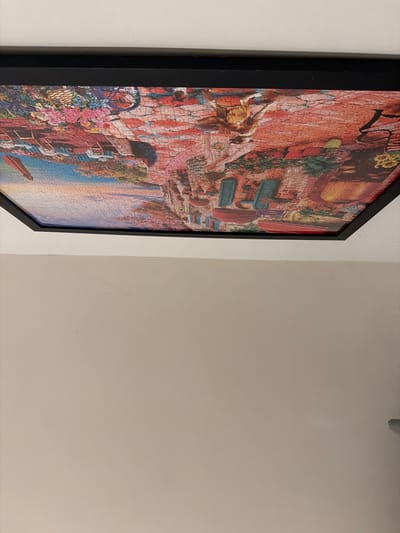 A framed jigsaw puzzle depicting a colorful scene of flowers, buildings, and a bridge is hanging on a plain, off-white wall. The puzzle is predominantly red, pink, and blue, with green accents. The style appears to be illustrative or painterly. The puzzle is mounted within a thin black frame. The image is taken from a low angle, looking upwards at the framed puzzle. There is no visible information or context to suggest a specific location in Tewkesbury, United Kingdom.