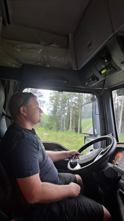 A man is driving a truck on a road surrounded by a forest. The man is wearing a black t-shirt and shorts. He has sunglasses on his head. The truck's interior is visible, including the steering wheel, dashboard, and a radio. The weather appears to be overcast. The image was taken in the village of Kabachigurt, Russia.